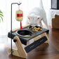 White cat eating from the double pet bowl stand with a hanging rod and a woven lantern ornament on a wooden floor