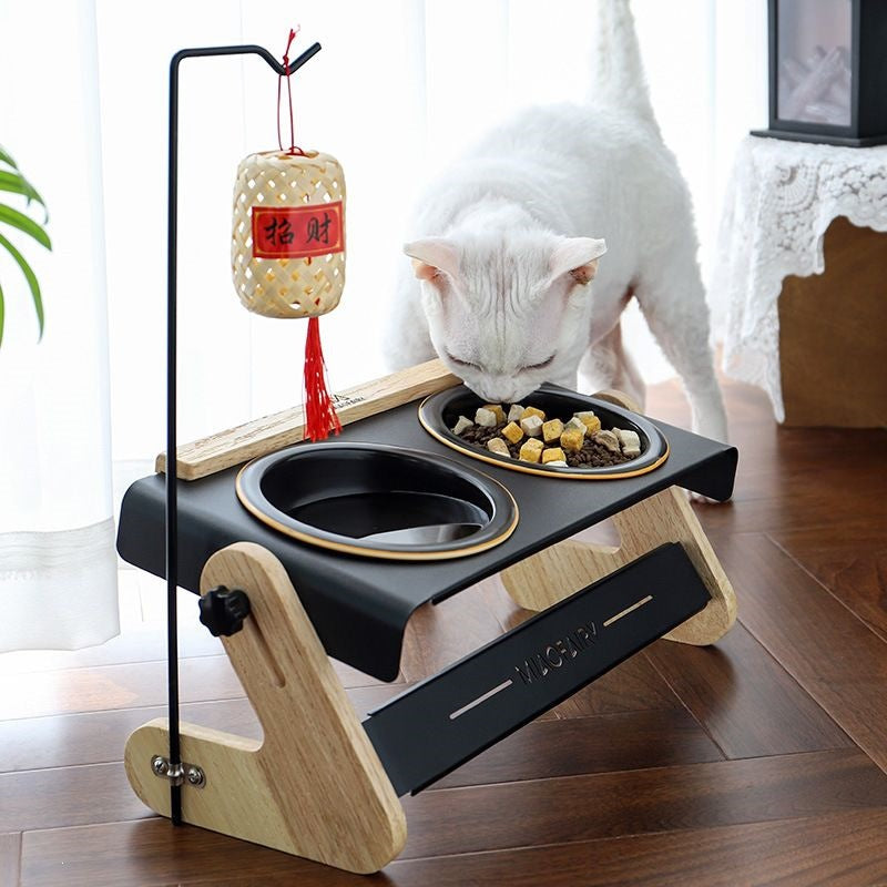 White cat eating from the double pet bowl stand with a hanging rod and a woven lantern ornament on a wooden floor
