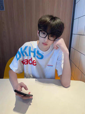 Young man with glasses seated at a table in a loose white t-shirt with large blue and red letters and a small sleeve print