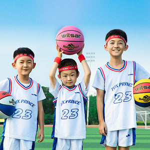 Three children in basketball jerseys holding a kids basketball together outdoors on a field