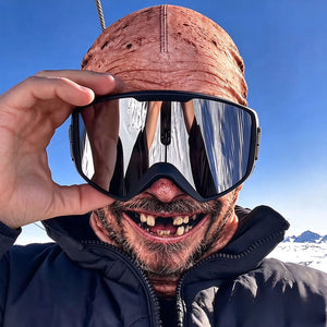Man wearing a printed elder face balaclava with silver ski goggles lifted in his hand under a clear blue sky