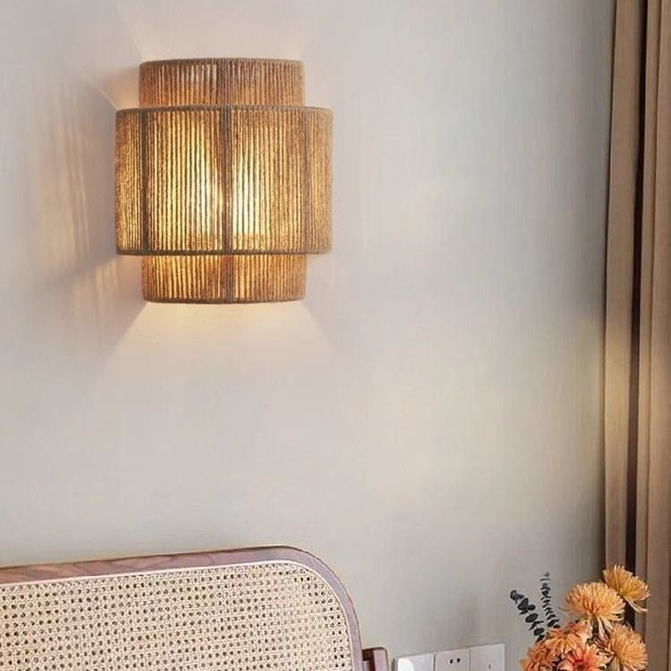 Hemp rope wall lamp glowing warm white on a wall behind a cane chair back, curtain and orange flowers