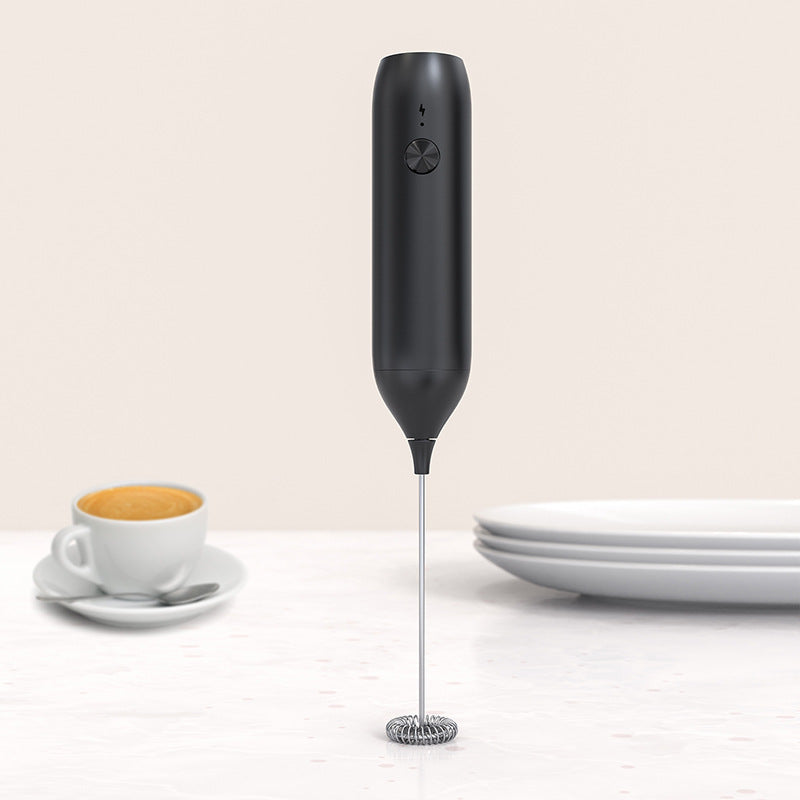 Black rechargeable milk frother with spring whisk standing on a counter beside a cup of coffee