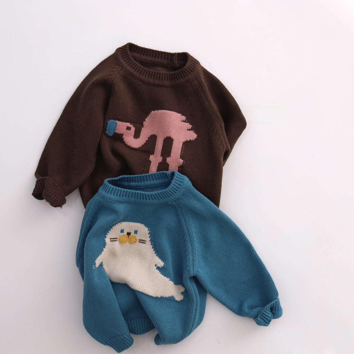 Brown flamingo knit sweater laid above a blue seal knit sweater on a pale background