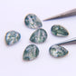 Six moss agate pears on white with steel tweezers, faceted crowns catching the light