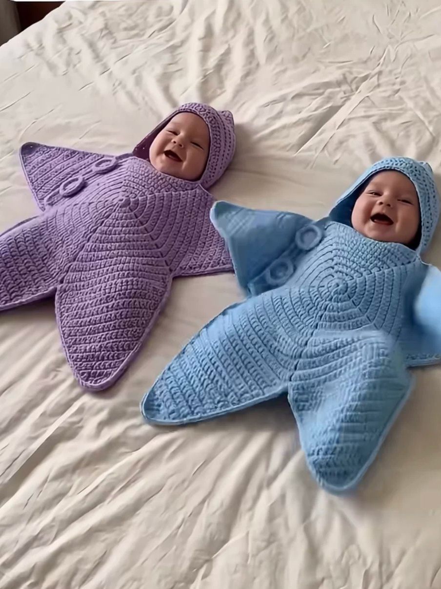 Purple and blue crochet star hooded baby buntings worn by two babies lying on a bed