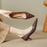 Woman holding a brown pointed toe pump above the other pump on her foot, low flared heel, on a woven rug
