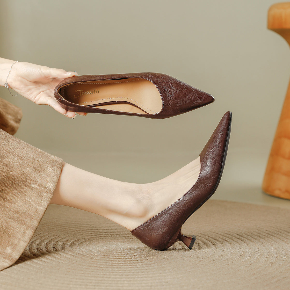 Woman holding a brown pointed toe pump above the other pump on her foot, low flared heel, on a woven rug