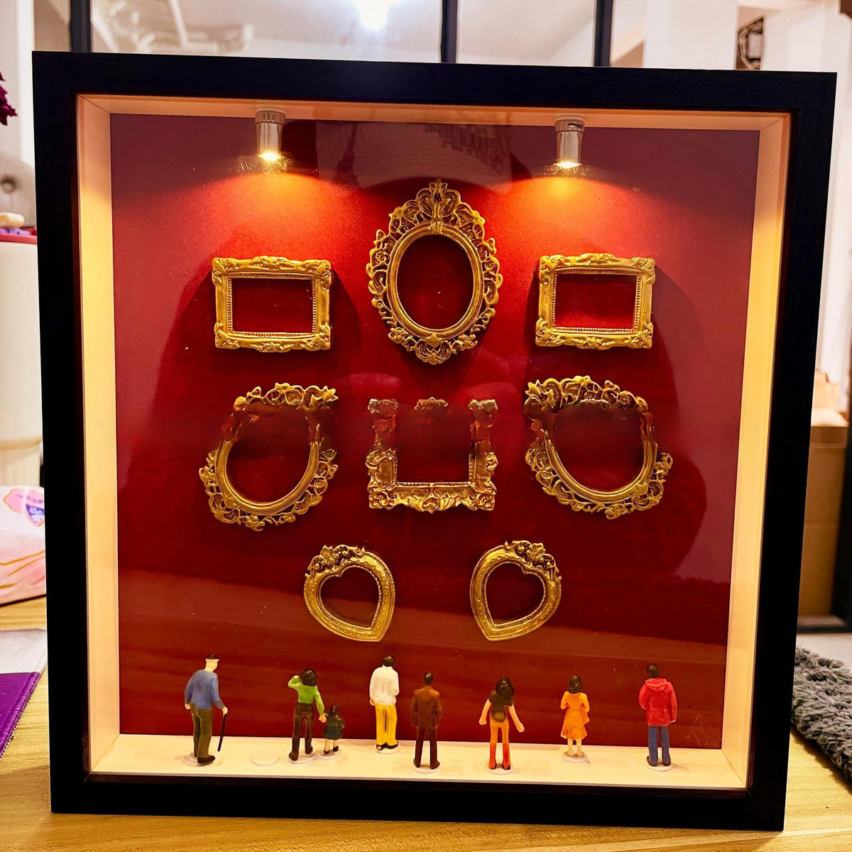 Black frame red backdrop shadow box lit by two spotlights with empty gold frames and small figurines on the ledge