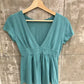 Teal short-sleeve deep V-neck top with a smooth waist band on a wooden hanger against a concrete wall