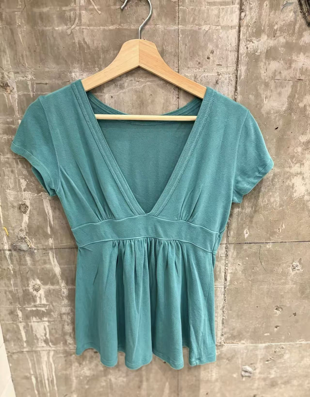 Teal short-sleeve deep V-neck top with a smooth waist band on a wooden hanger against a concrete wall