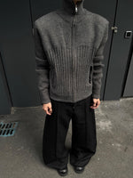 Man in a charcoal gray zip front ribbed knit cardigan and wide black trousers standing against a dark wall