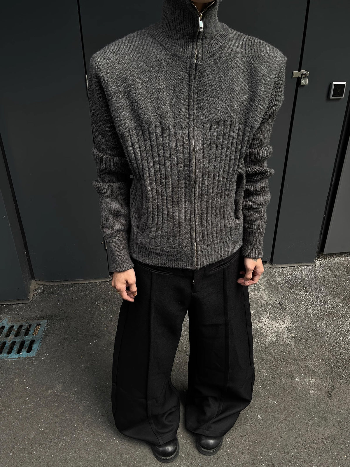 Man in a charcoal gray zip front ribbed knit cardigan and wide black trousers standing against a dark wall
