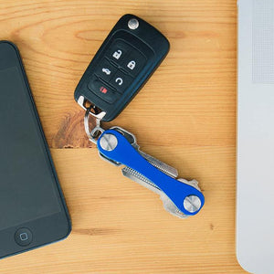 Blue aluminum key organizer clipped beside a black car key fob on a wooden desk