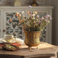 Gold iron flowerpot with pierced teardrop pattern on a wooden table with flowers, a teacup and a framed picture
