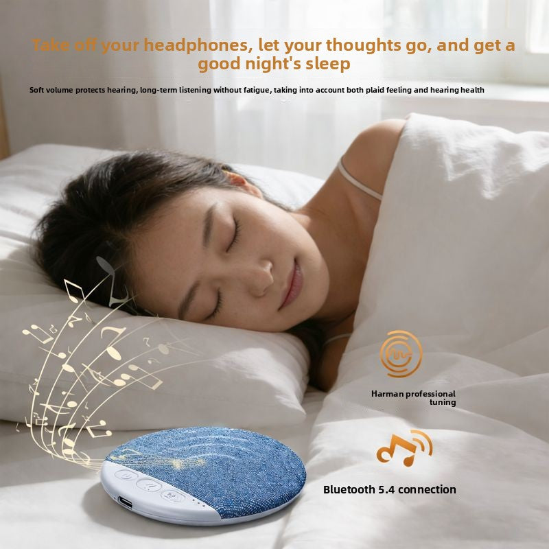 Grey fabric pillow speaker lying flat on a white sheet next to a pillow