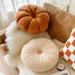 Orange pumpkin pillow and beige round pleated pillow with an orange button on a white throw beside a checkerboard pillow