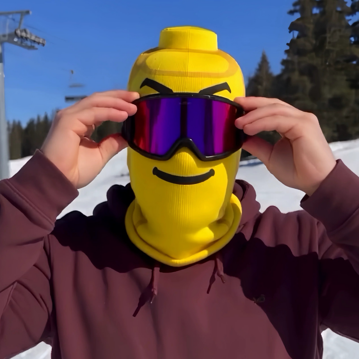 Person lifting purple ski goggles on a yellow block head printed face cover, mountain slope behind