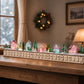 Pastel pink wooden advent calendar village with lit windows near a holiday wreath