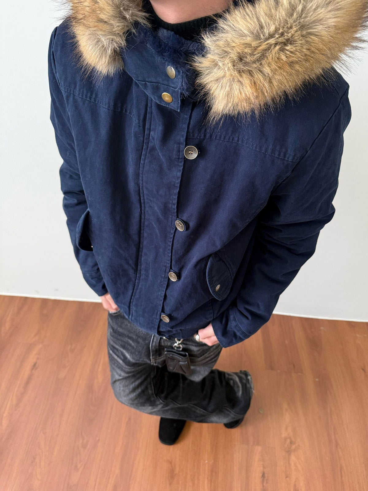 Navy blue washed hooded jacket with a vegan fur trimmed hood and metal snap front, worn with dark jeans on a wooden floor