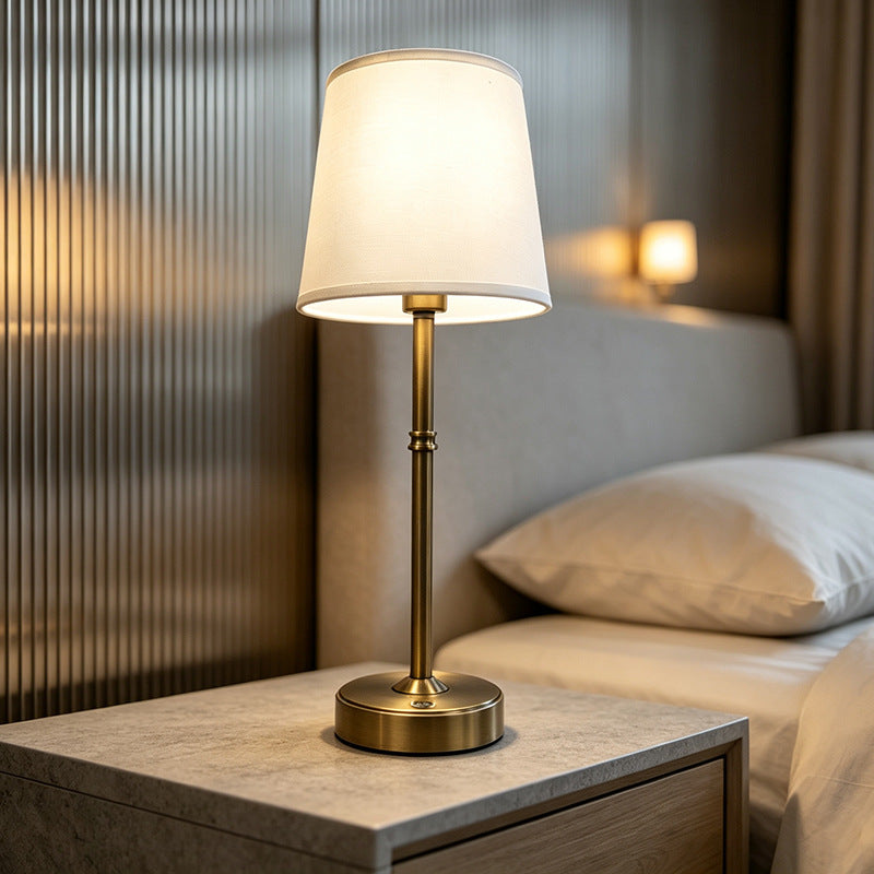 Lit gold metal table lamp with white fabric shade on a bedroom nightstand at night
