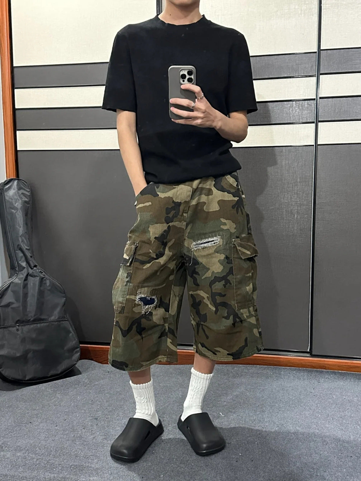 Model in a black tee and camo patchwork denim cargo shorts taking a mirror photo, hem below the knee