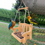 Natural wood bench shaped bird feeder hanging from a branch with seeds on the tray and two painted birds beside it