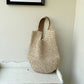 Slouchy paper straw shoulder bag standing on a chipped white side table, tan straps upright