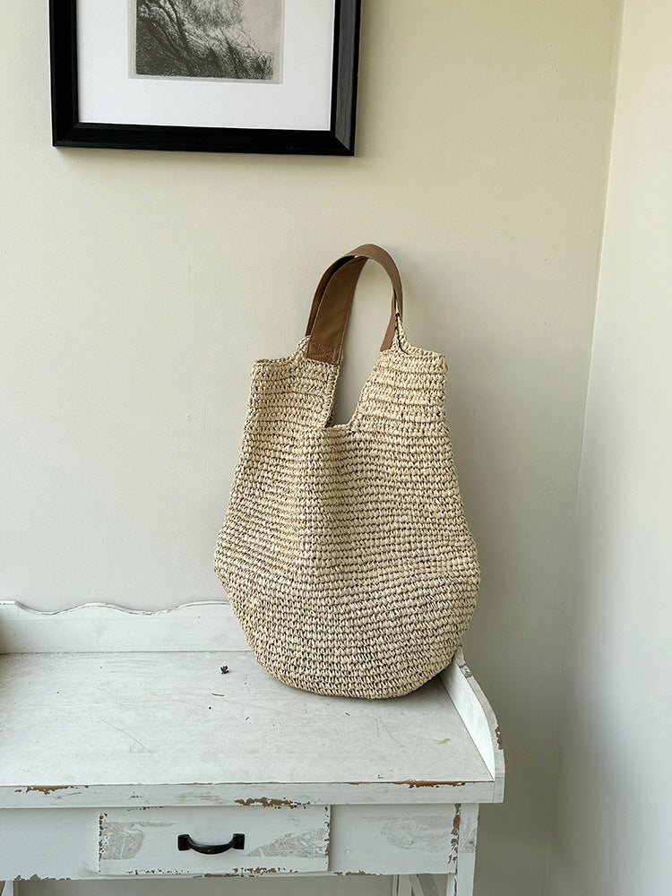Slouchy paper straw shoulder bag standing on a chipped white side table, tan straps upright