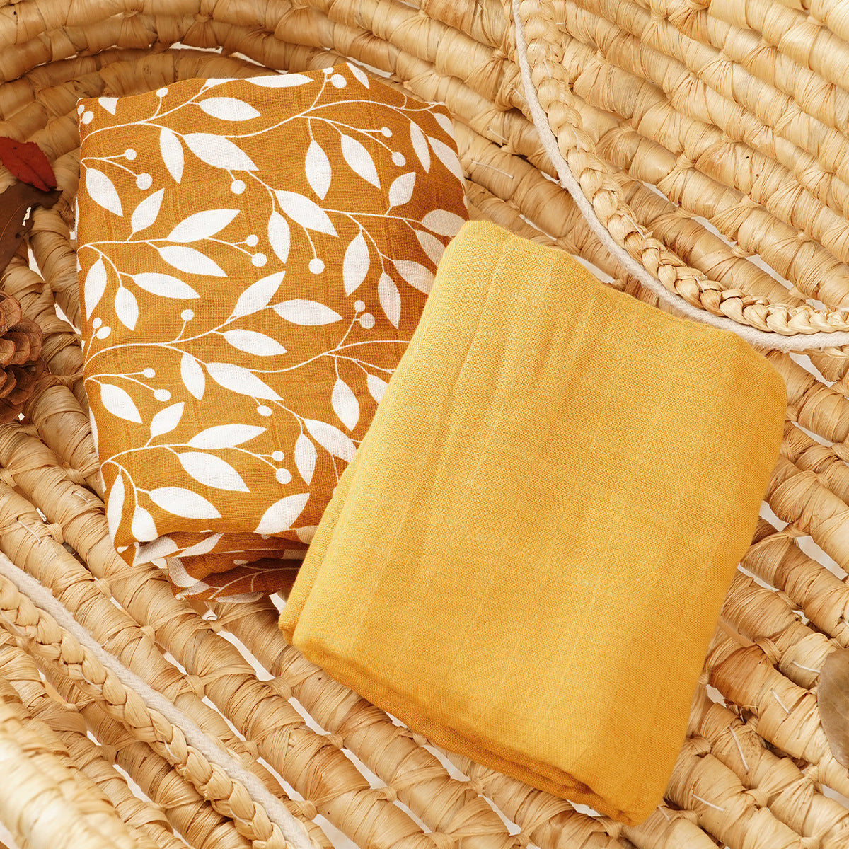 Two folded cotton muslin baby blankets, one mustard botanical print and one solid mustard, in a basket