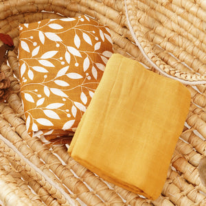 Two folded cotton muslin baby blankets, one mustard botanical print and one solid mustard, in a basket
