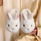 Pair of white bunny plush slippers with pink lined upright ears held in a hand against cream fabric, view 2 of 3