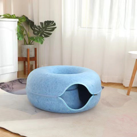 Cat peeking out of the tunnel opening of a beige felt donut bed on a floor