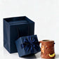 Red orange ceramic mug with a yellow crescent hold beside a navy blue gift box with a satin bow and a gift bag