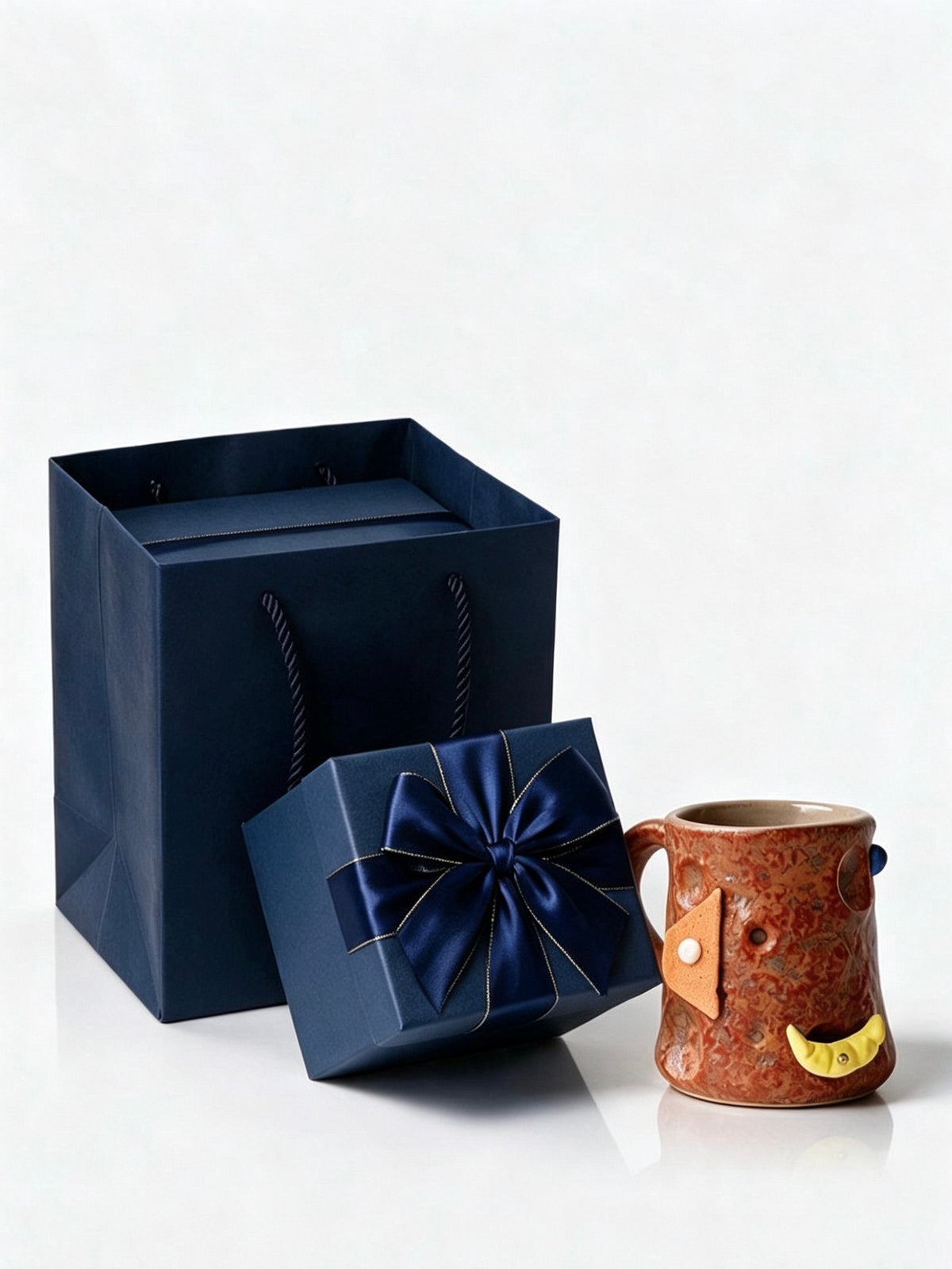 Red orange ceramic mug with a yellow crescent hold beside a navy blue gift box with a satin bow and a gift bag