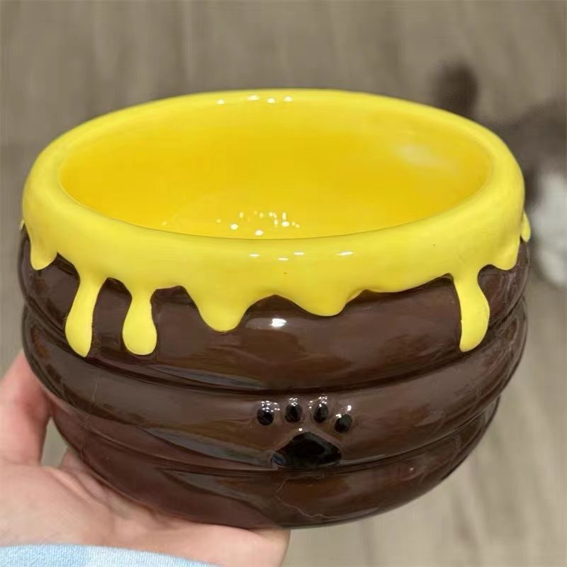 Hand holding a brown ceramic honey jar bowl with yellow drips and a paw print, close view