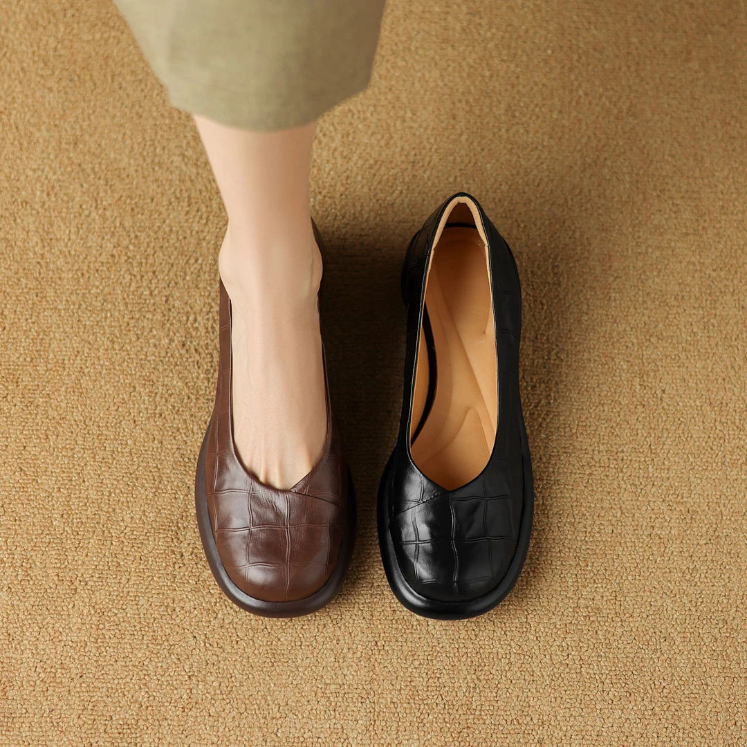 Brown and black embossed leather block heel slip-on shoes on a woven carpet beside a woman's foot