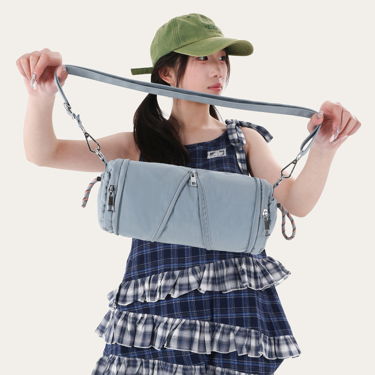 Woman holding the long strap of a dusty blue cylindrical nylon bag with silver hooks and orange cord loops