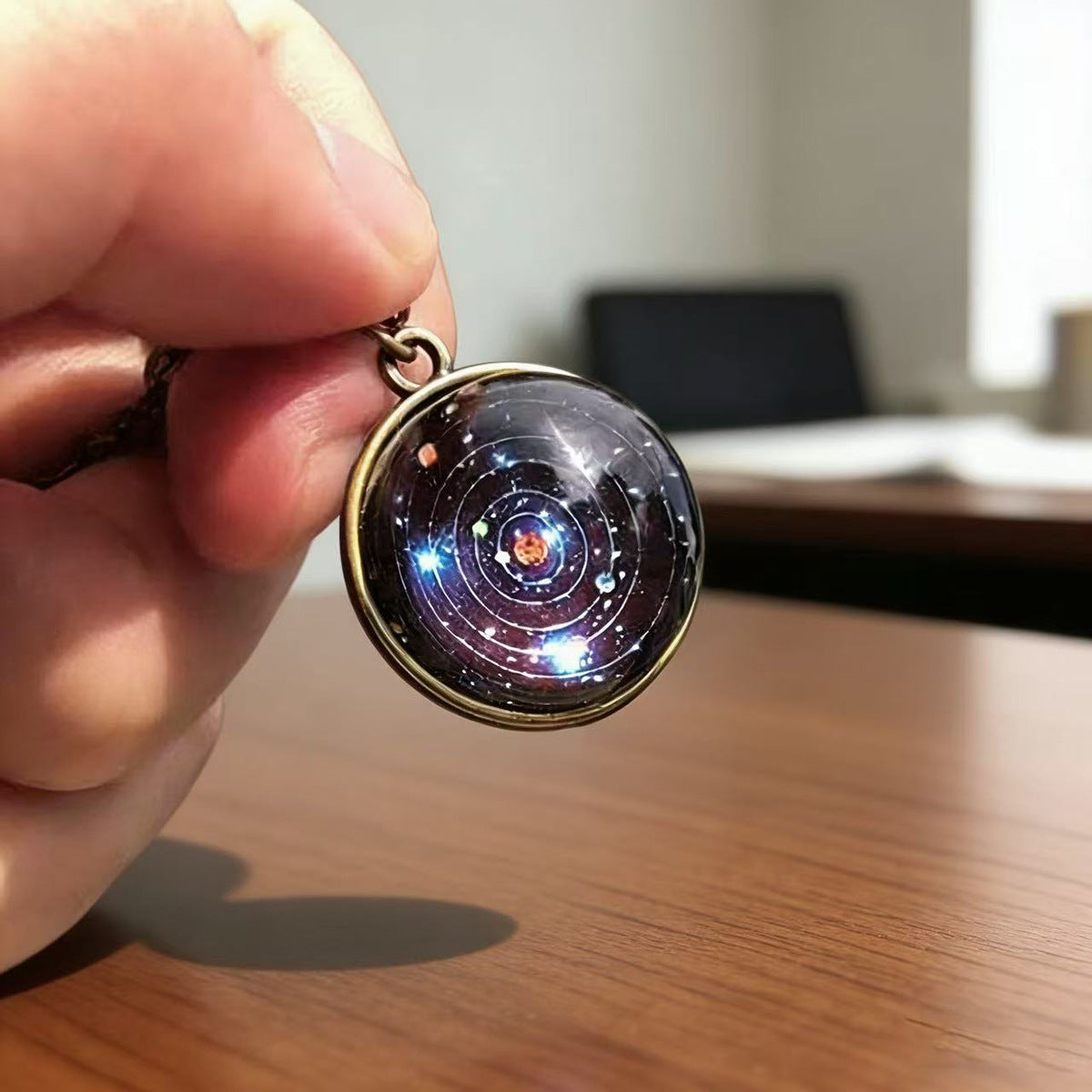 Glass solar system pendant held above a wooden desk, bronze tone ring and fine chain visible
