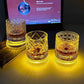 Three whiskey tumblers with different cut patterns glowing amber on a desk in a dark room