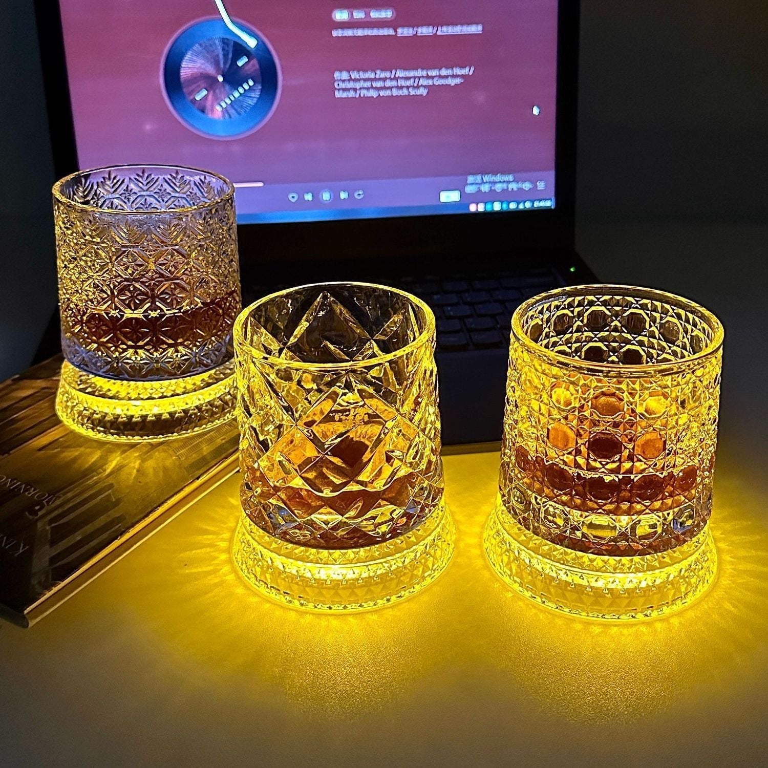 Three whiskey tumblers with different cut patterns glowing amber on a desk in a dark room