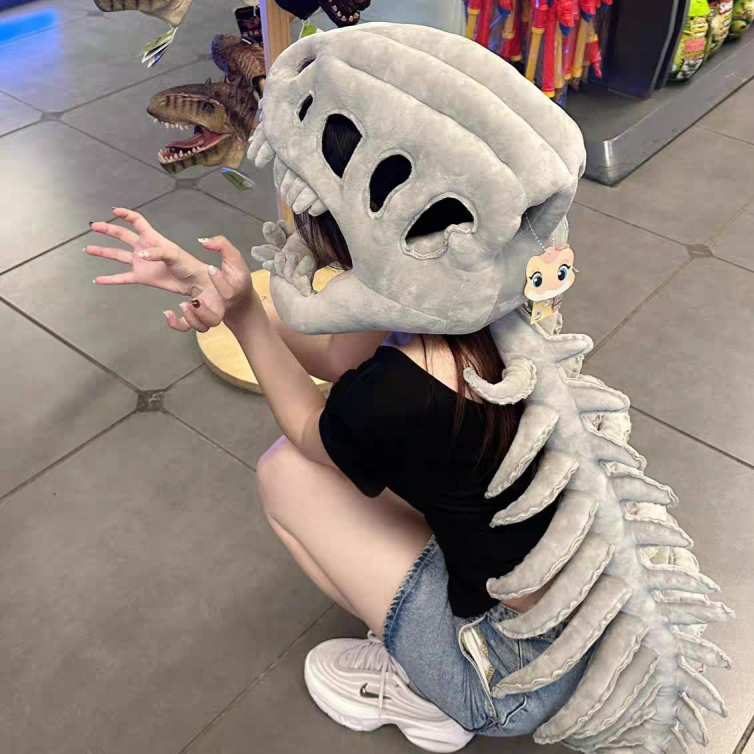 Grey plush dinosaur skeleton head mask worn while kneeling, spine trailing along the floor