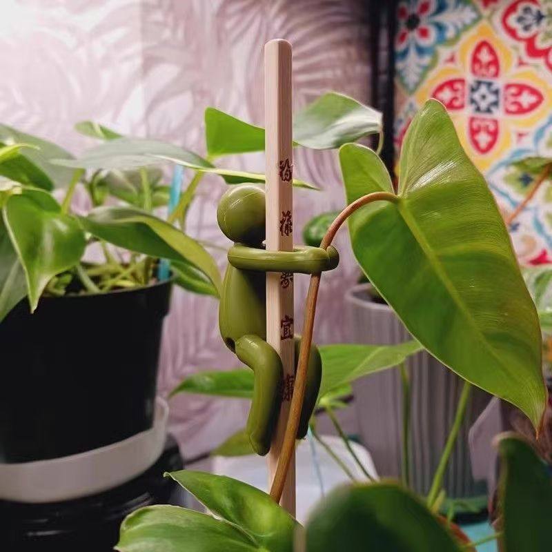 Olive green figure clinging with both arms to a plant stem and a wooden stake in a potted plant