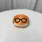 Orange resin frog earbud case wearing round black spectacles, closed, on a white cloth