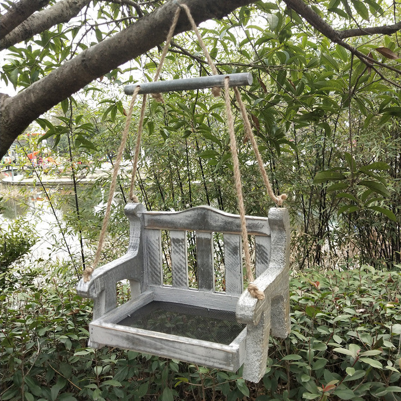 Weathered gray wooden bench bird feeder with a mesh floor and a gray crossbar hanging in a green garden