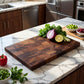 Walnut cutting board on a marble kitchen counter with fresh herbs, red onion, and lime