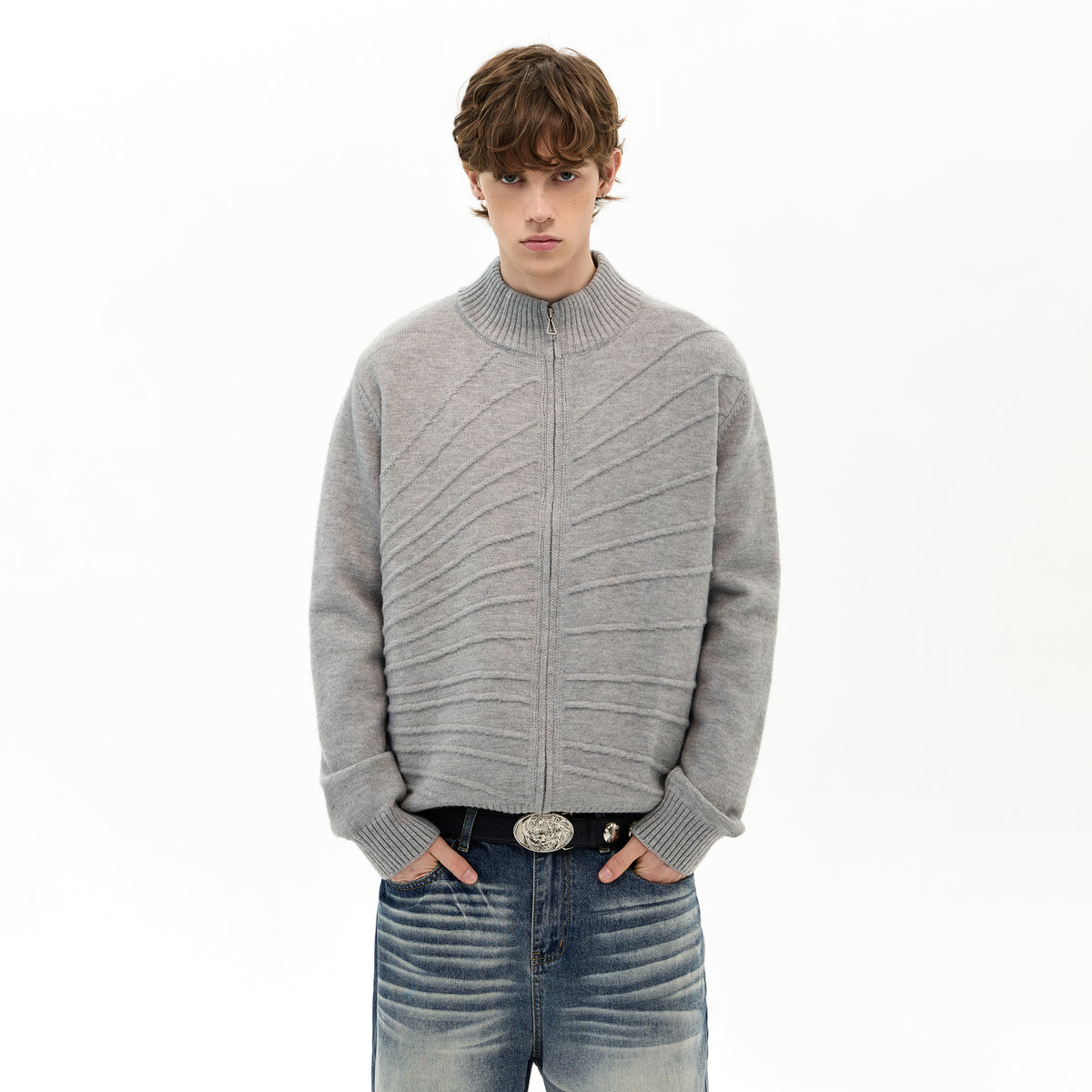 Model in a gray ribbed zip-up sweater and jeans with a belt buckle, standing against a white background