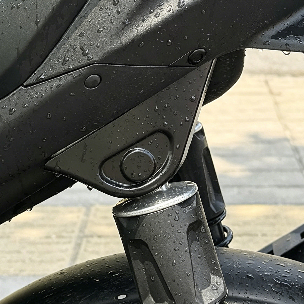 Black plastic cover over a motorcycle rear shock absorber mount, beaded with rain drops