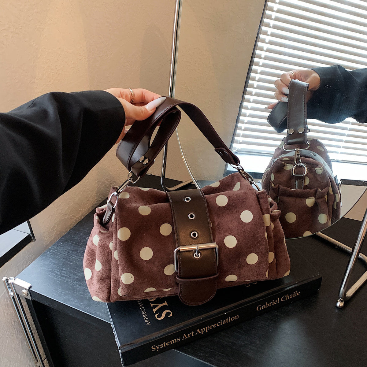 Coffee brown polka dot shoulder bag with wide front buckle band held by its short strap beside a mirror