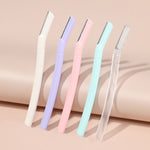 Five eyebrow razors in white, lilac purple, pink, mint green and frosted transparent leaning on a peach backdrop
